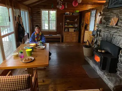 a woman sitting at a wooden table in a room with a fireplace at Private Lakefront Family Cottage with Tennis Court in Kinmount